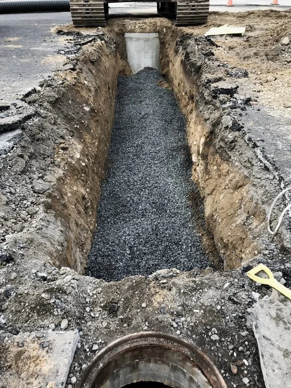 A residential construction site with an excavated sewage pipeline. The sewer line trench is filled with gravel, with an excavator parked at the top edge of the trench.