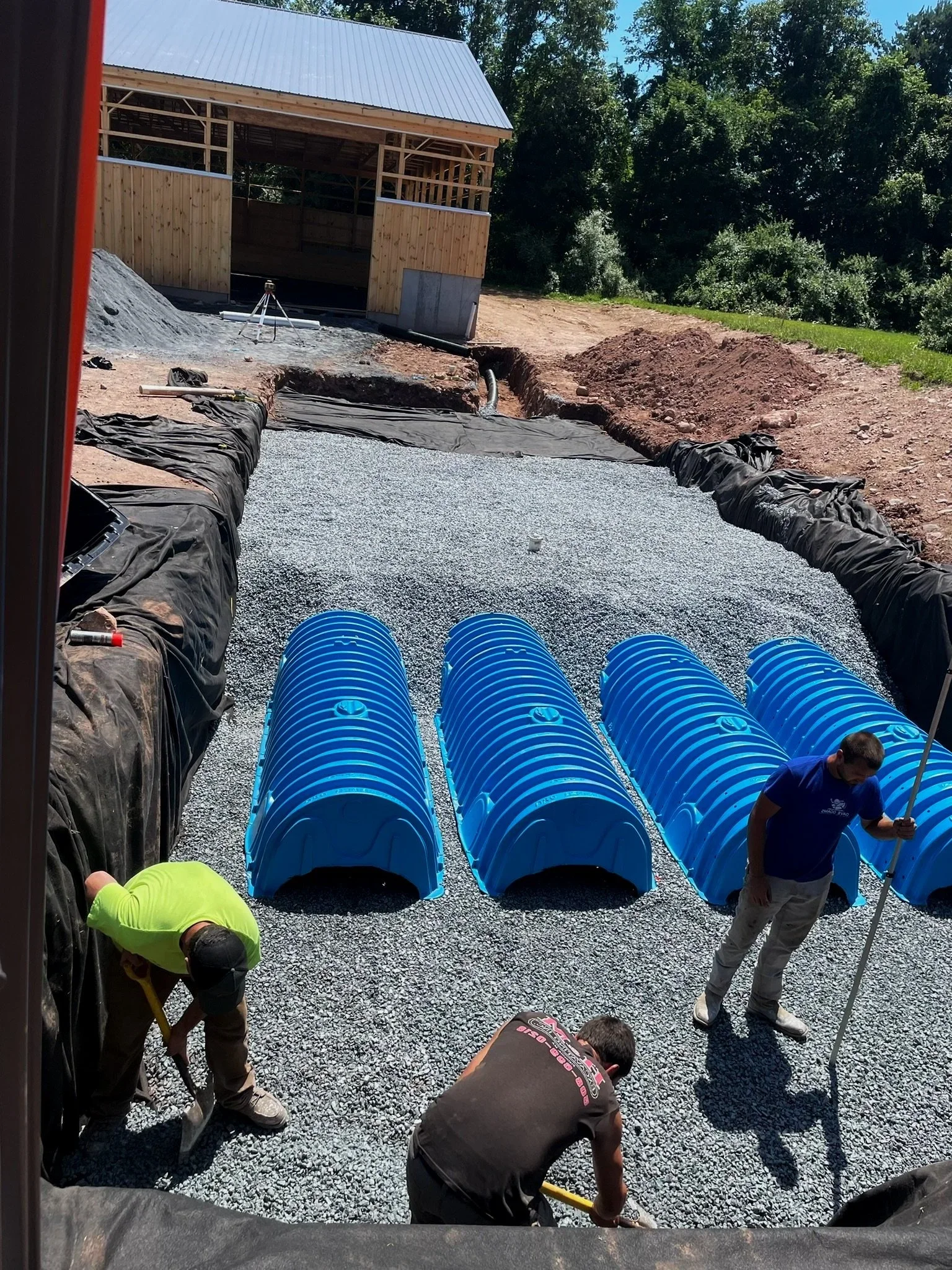Excavation workers are installing a residential waterline pathway. Blue curved piping is in a gravel-filled pit with a wooden shed visible in the background.