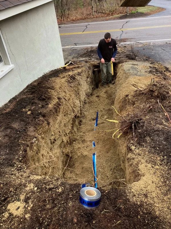 An excavation specialist in a black shirt and gray pants is measuring a freshly dug waterline trench outside a white home with a yellow level tool. Blue waterline pipes run through the center of the trench, surrounded by exposed roots and soil.