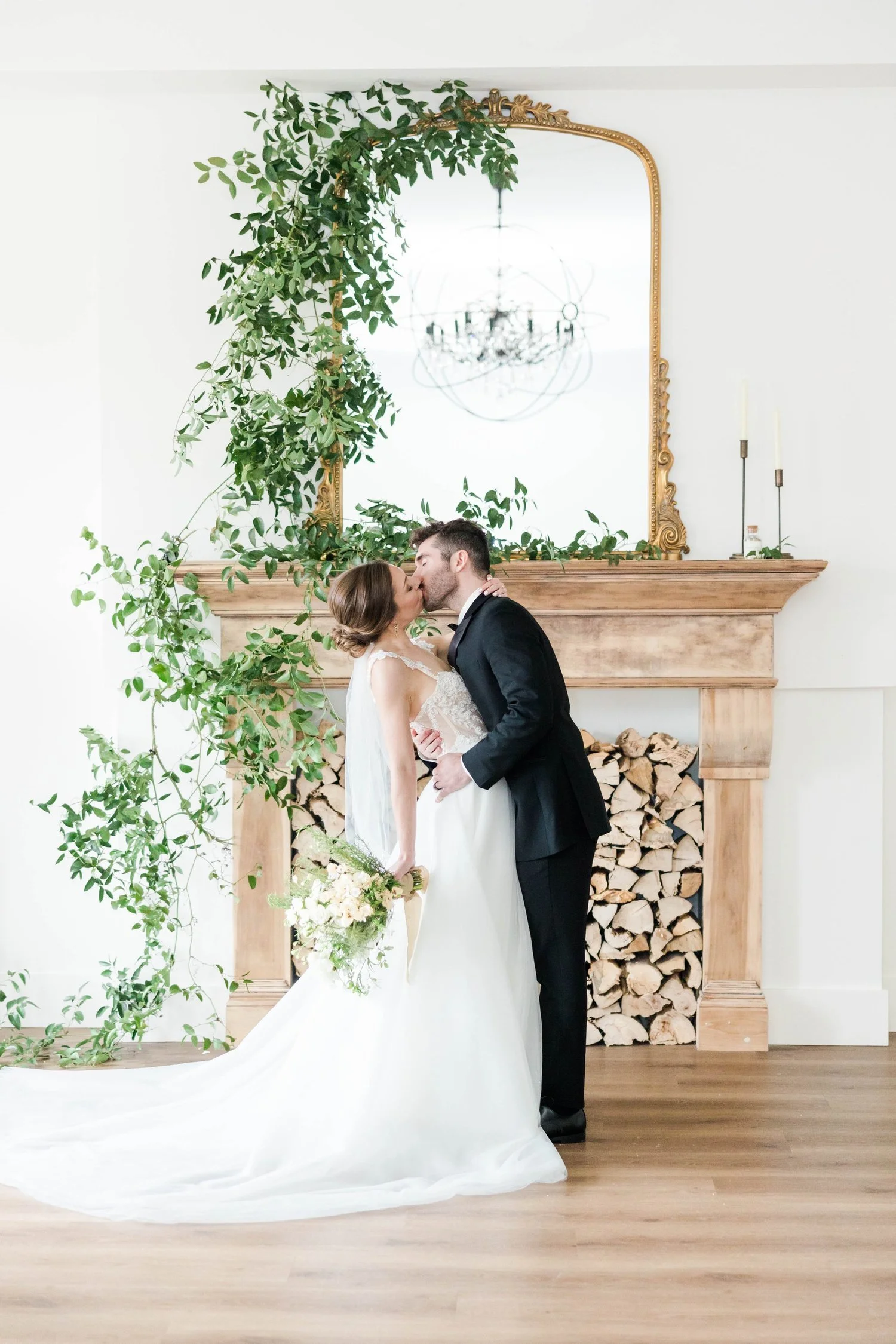 Couple kissing in front of a wood mantel draped in trailing greenery with a gold mirror above, at The Broadway Avenue