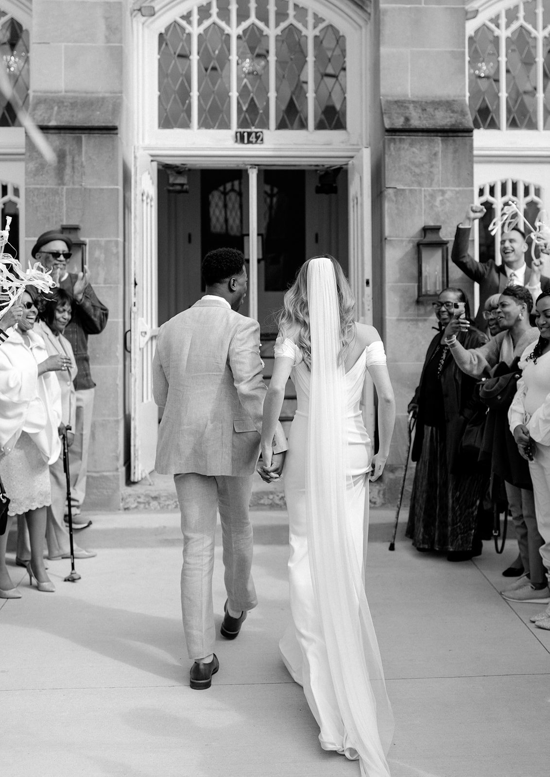 Black-and-white photo of a couple walking out through arched church doors as guests wave ribbon wands, at The Broadway Avenue