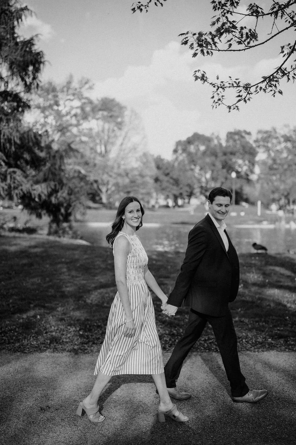 Black and white photo of a couple holding hands and walking beside a park pond, a relaxed setting among park engagement photoshoot locations.