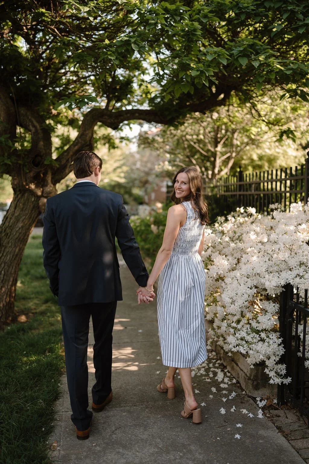 Woman smiling back over her shoulder while holding her fiancé's hand on a sidewalk lined with white azaleas.