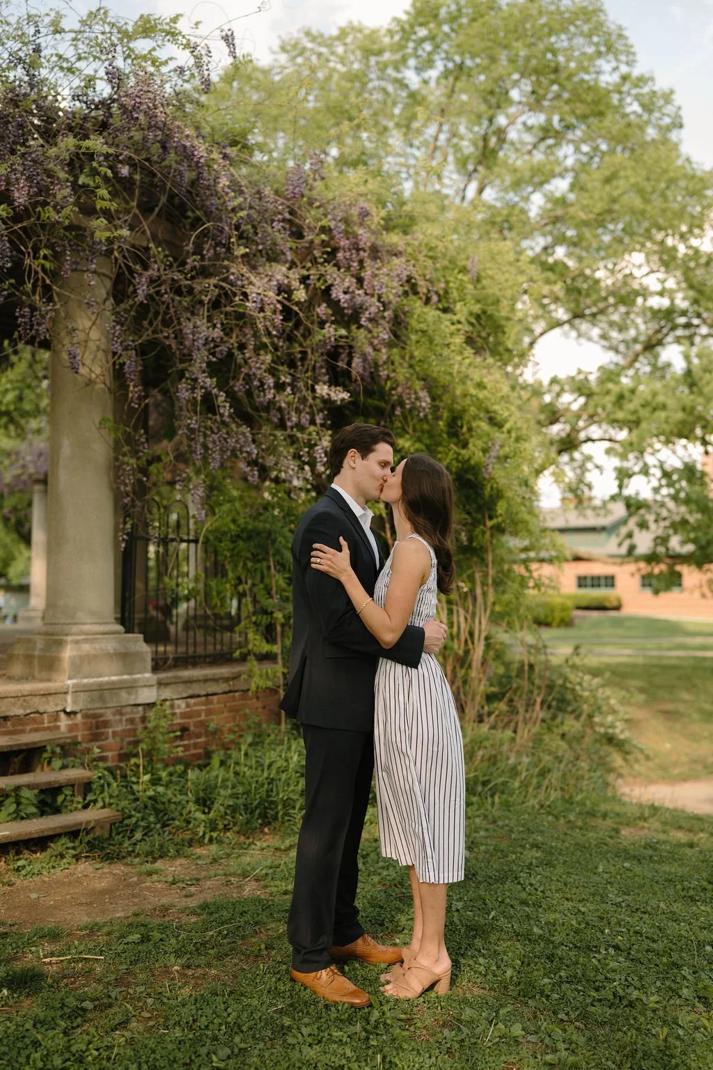 Couple sharing a kiss on the grass beside a stone column overflowing with purple wisteria.