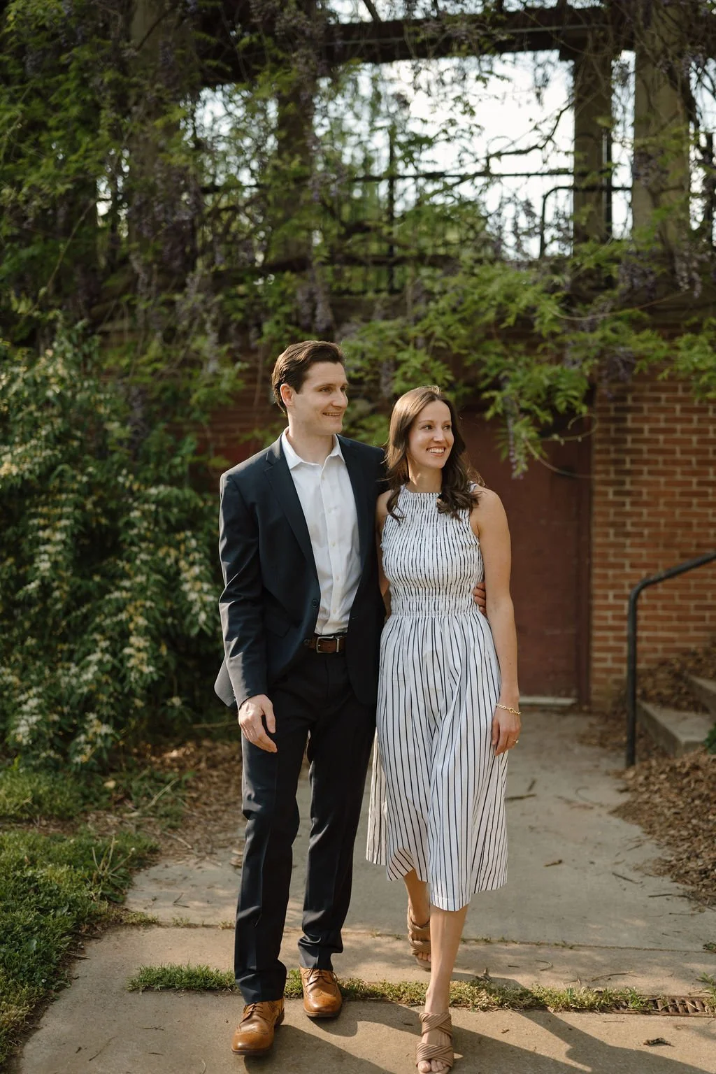Couple walking side by side past wisteria vines and a red brick wall, a cozy corner among spring engagement photoshoot locations.