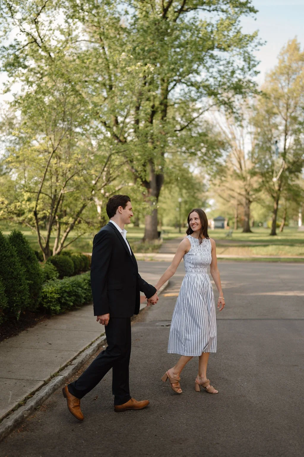 Woman leading her fiancé by the hand across a quiet tree-lined street in a striped midi dress.