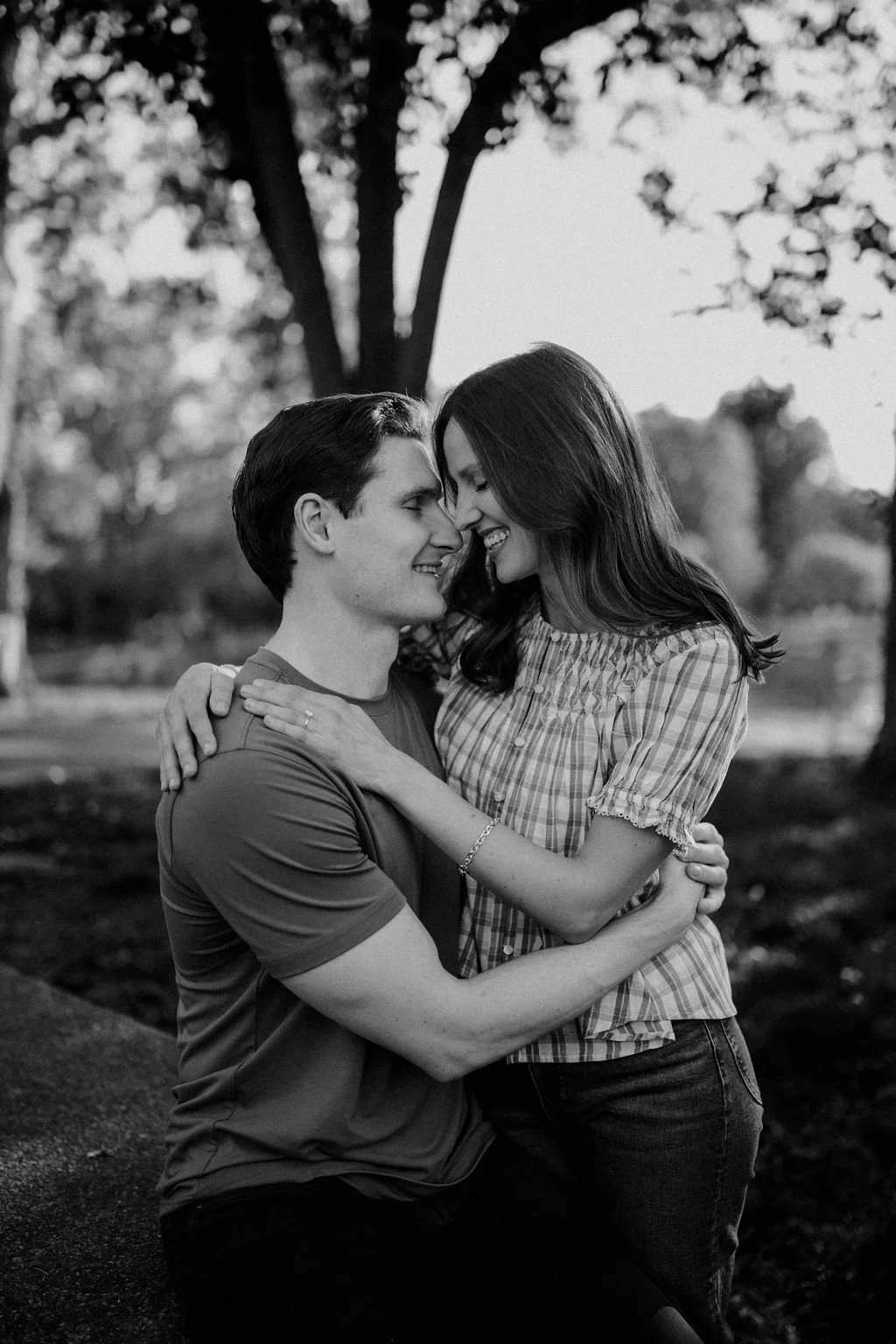 Black and white photo of a couple nose to nose and laughing in a warm embrace under the trees.