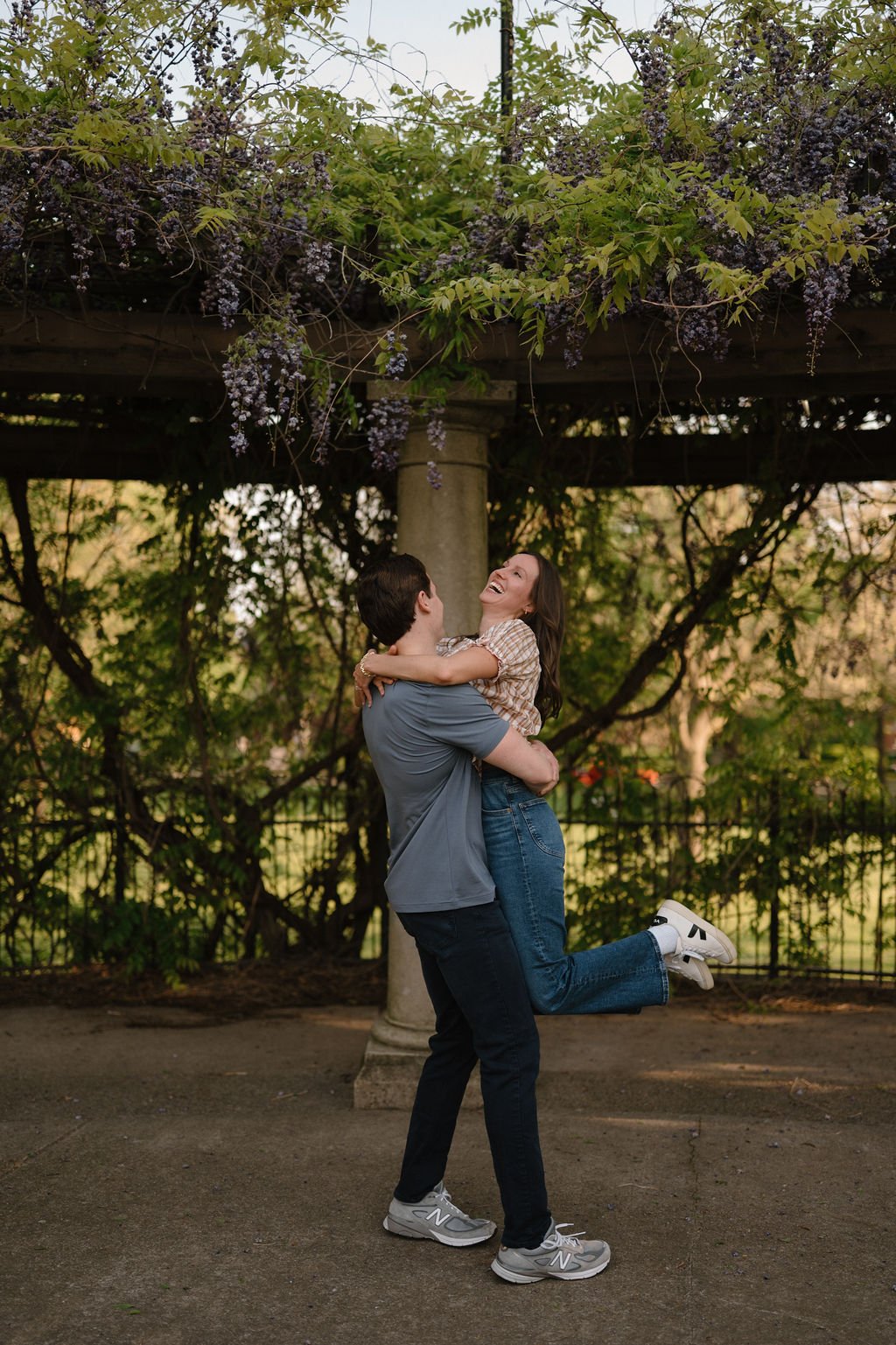 Man lifting his laughing fiancée off the ground under a wisteria pergola, proof that playful moments shine at outdoor engagement photoshoot locations.