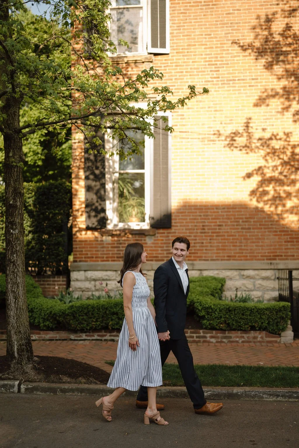 Couple holding hands and walking past a sunlit brick house with black shutters, a charming idea for historic neighborhood engagement photoshoot locations.