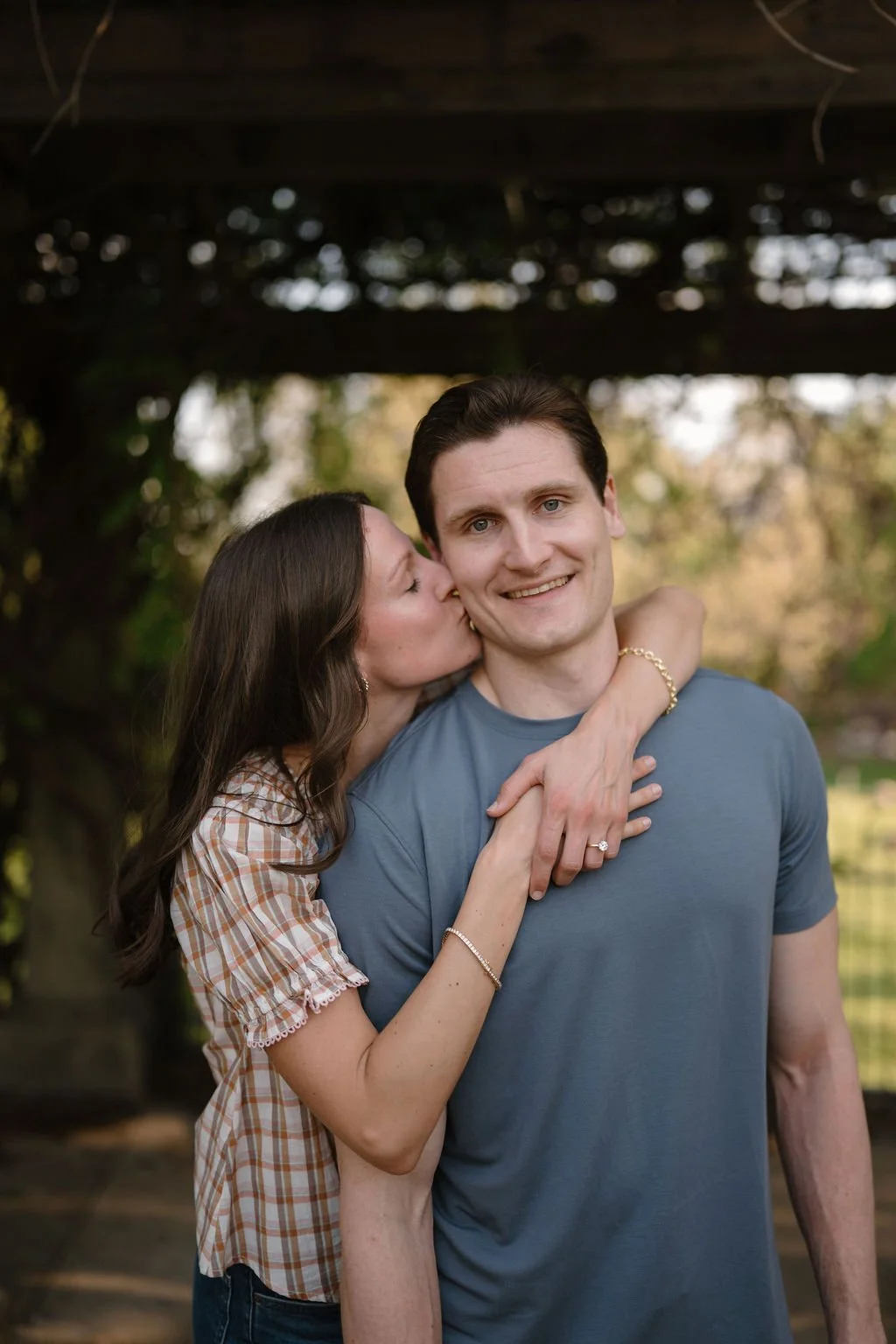 Woman kissing her fiancé on the cheek as he smiles at the camera under a garden pergola.