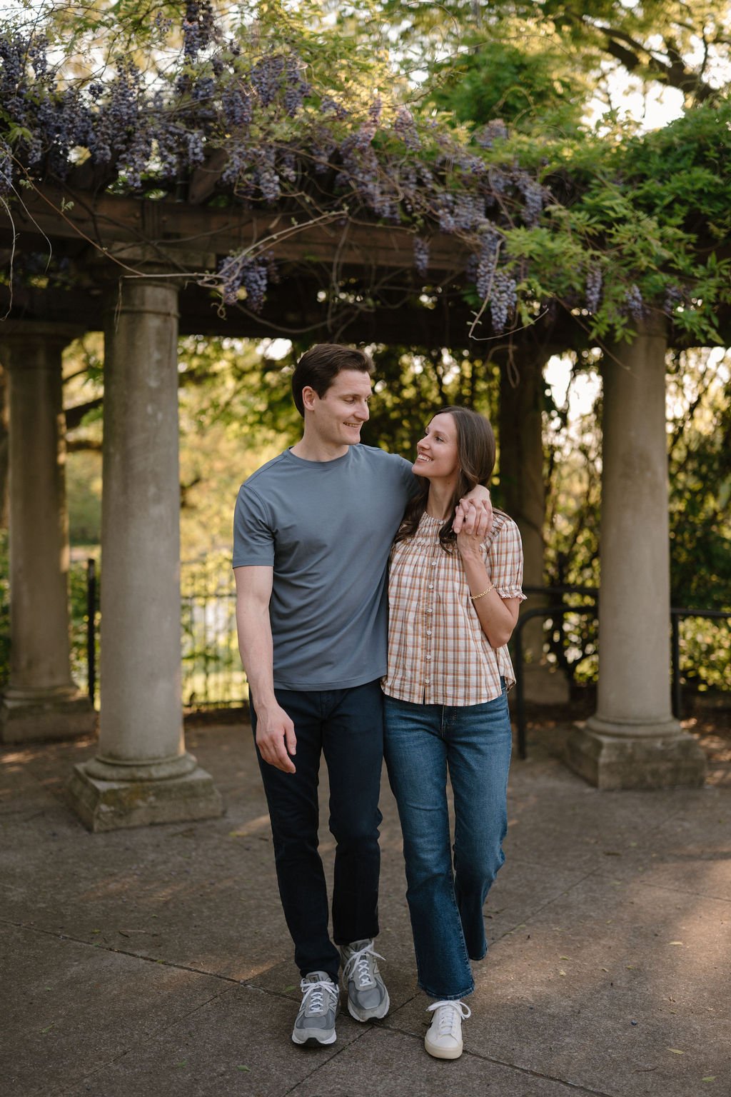 Couple walking arm in arm beneath a stone-column pergola draped in purple wisteria, a romantic choice for garden engagement photoshoot locations.