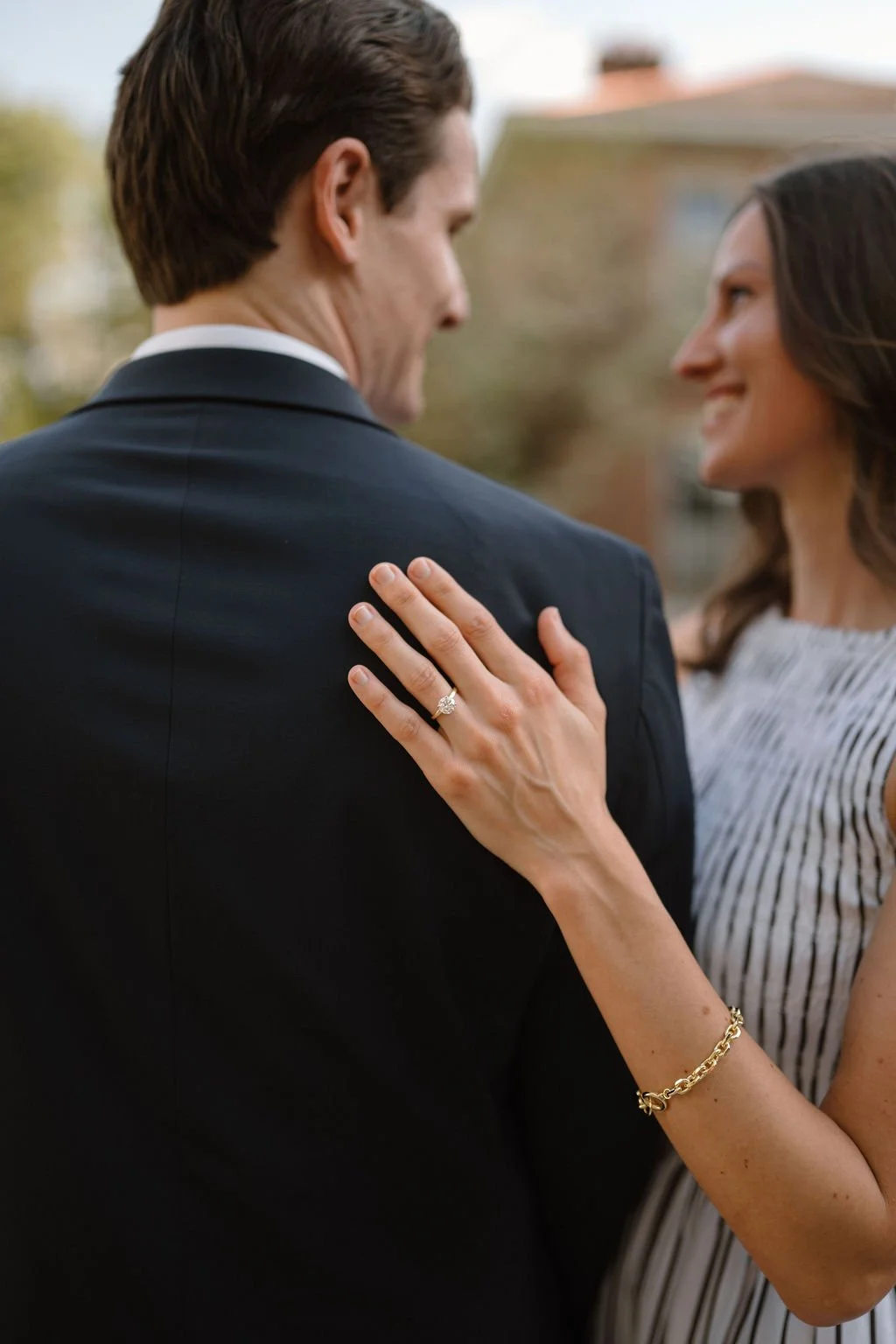 Close-up of a solitaire engagement ring and gold chain bracelet on a hand resting on a navy suit jacket, a detail shot that works at any engagement photoshoot location.