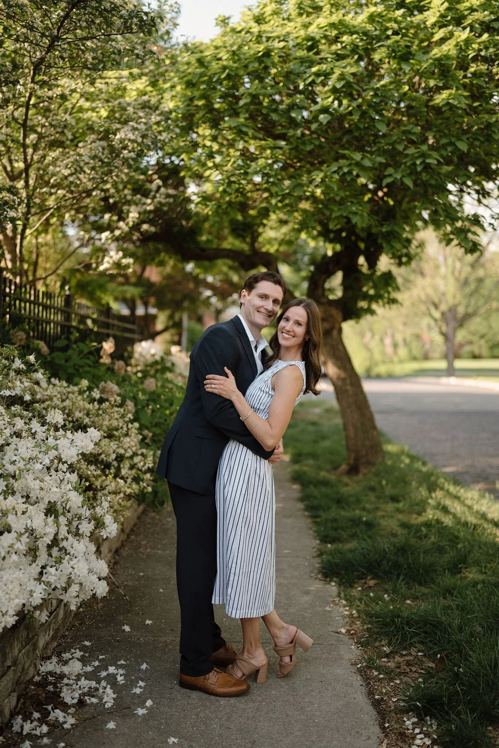 Couple hugging and smiling on a sidewalk beside blooming white azaleas under a shady tree.