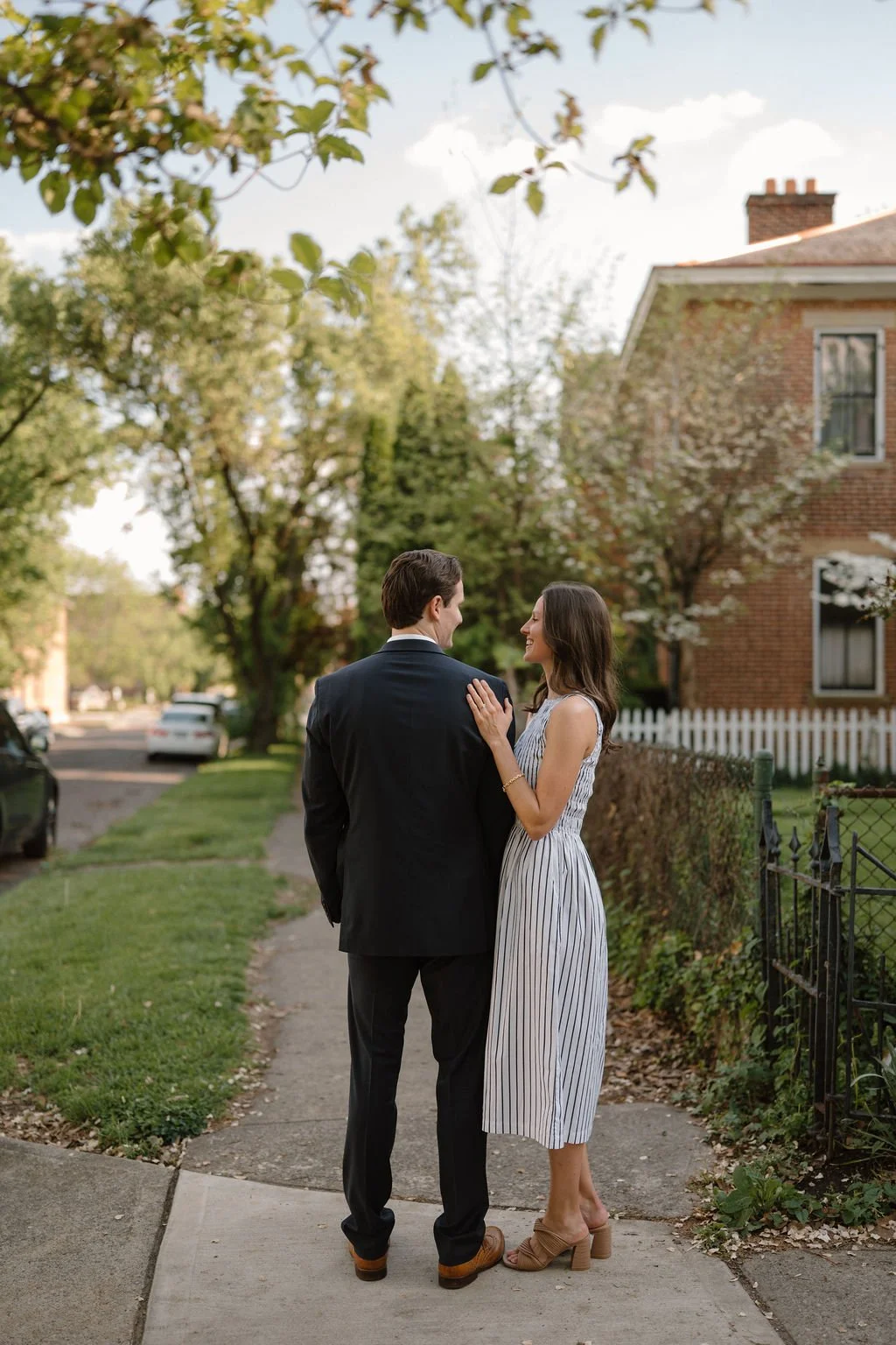 Couple standing on a tree-lined sidewalk facing each other with her hand resting on his shoulder near a red brick home.