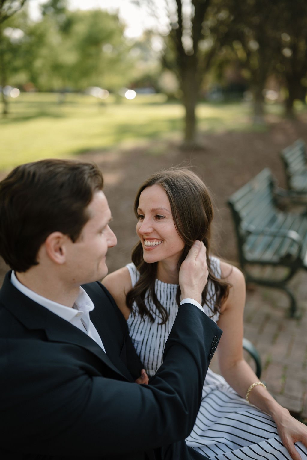 Man tucking his fiancée's hair behind her ear as she smiles at him on a park bench.