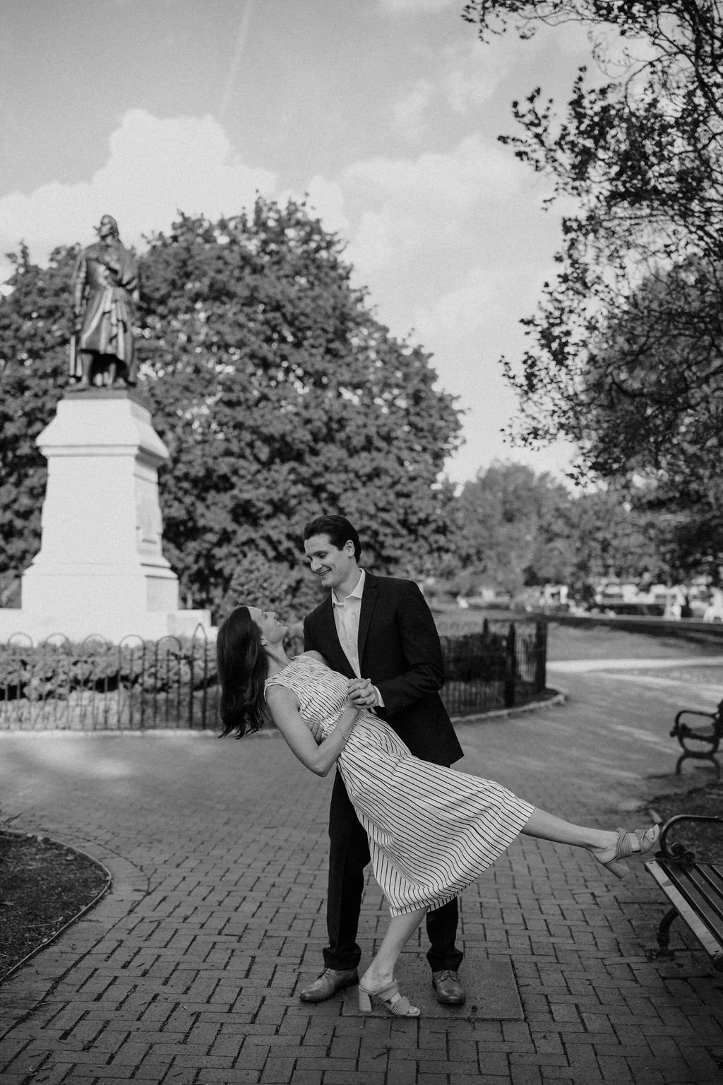 Black and white photo of a man dipping his fiancée in a striped dress beside a historic statue, one of the timeless city engagement photoshoot locations.