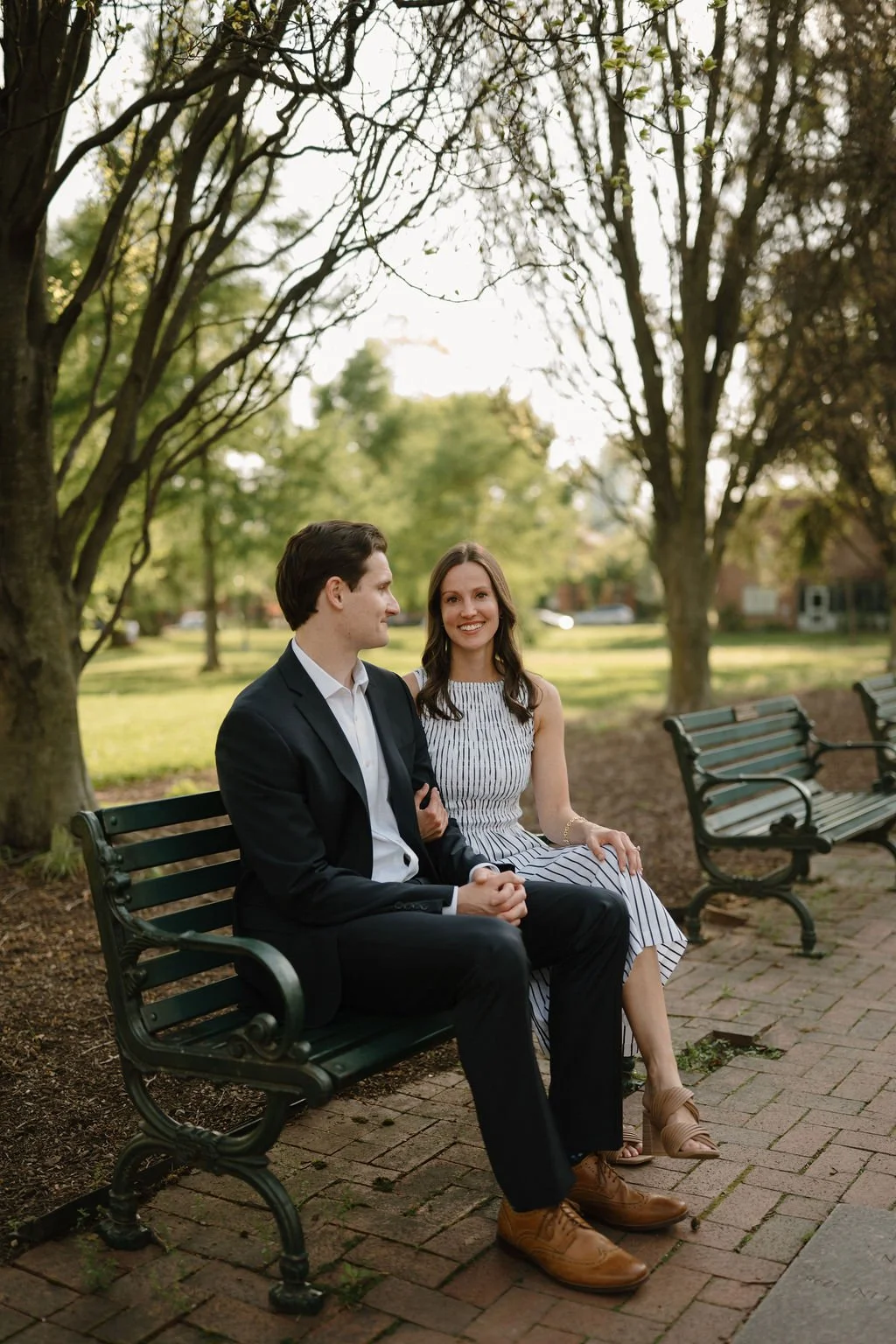 Couple sitting together on a green park bench under leafy trees, a classic pick for park engagement photoshoot locations.