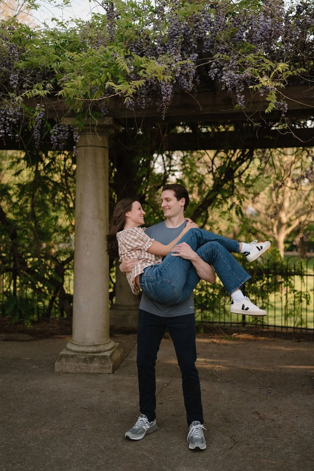 Man carrying his fiancée in his arms under a wisteria-covered pergola, a dreamy spot among spring engagement photoshoot locations.