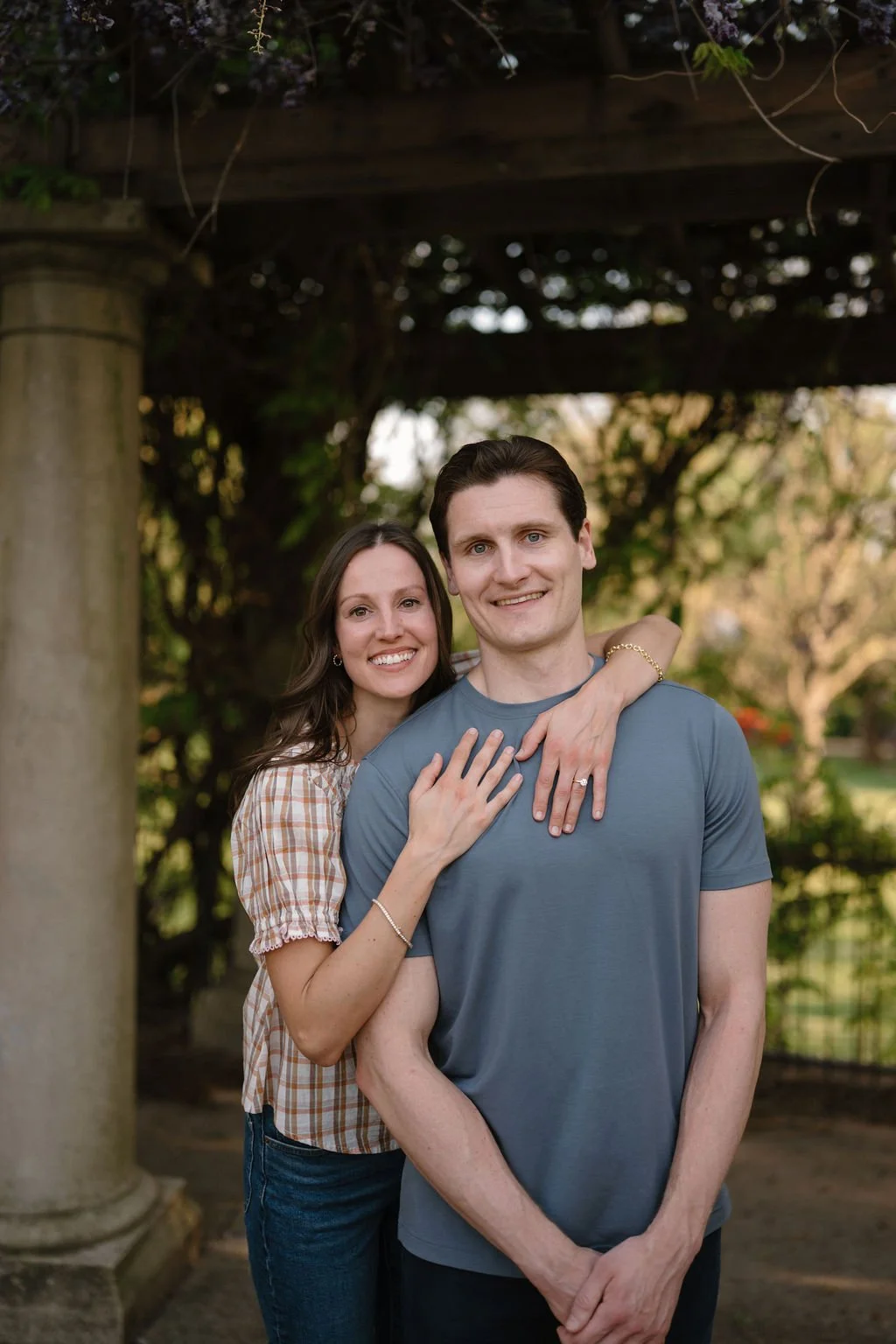 Smiling couple posing under a wooden pergola with her arms wrapped around his shoulders, showing off her engagement ring.