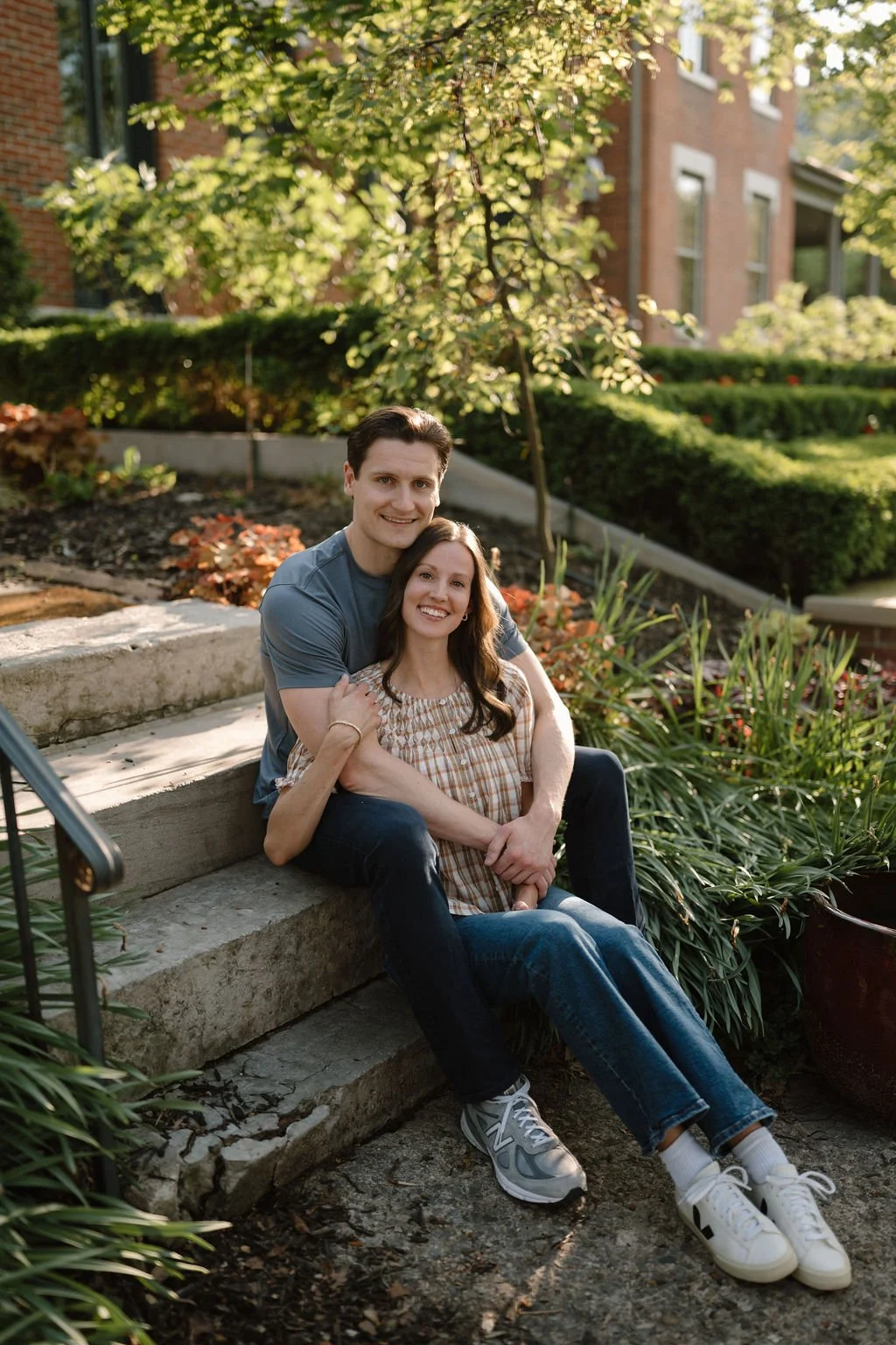 Couple sitting on stone garden steps, foreheads touching, surrounded by colorful plants and potted pottery.