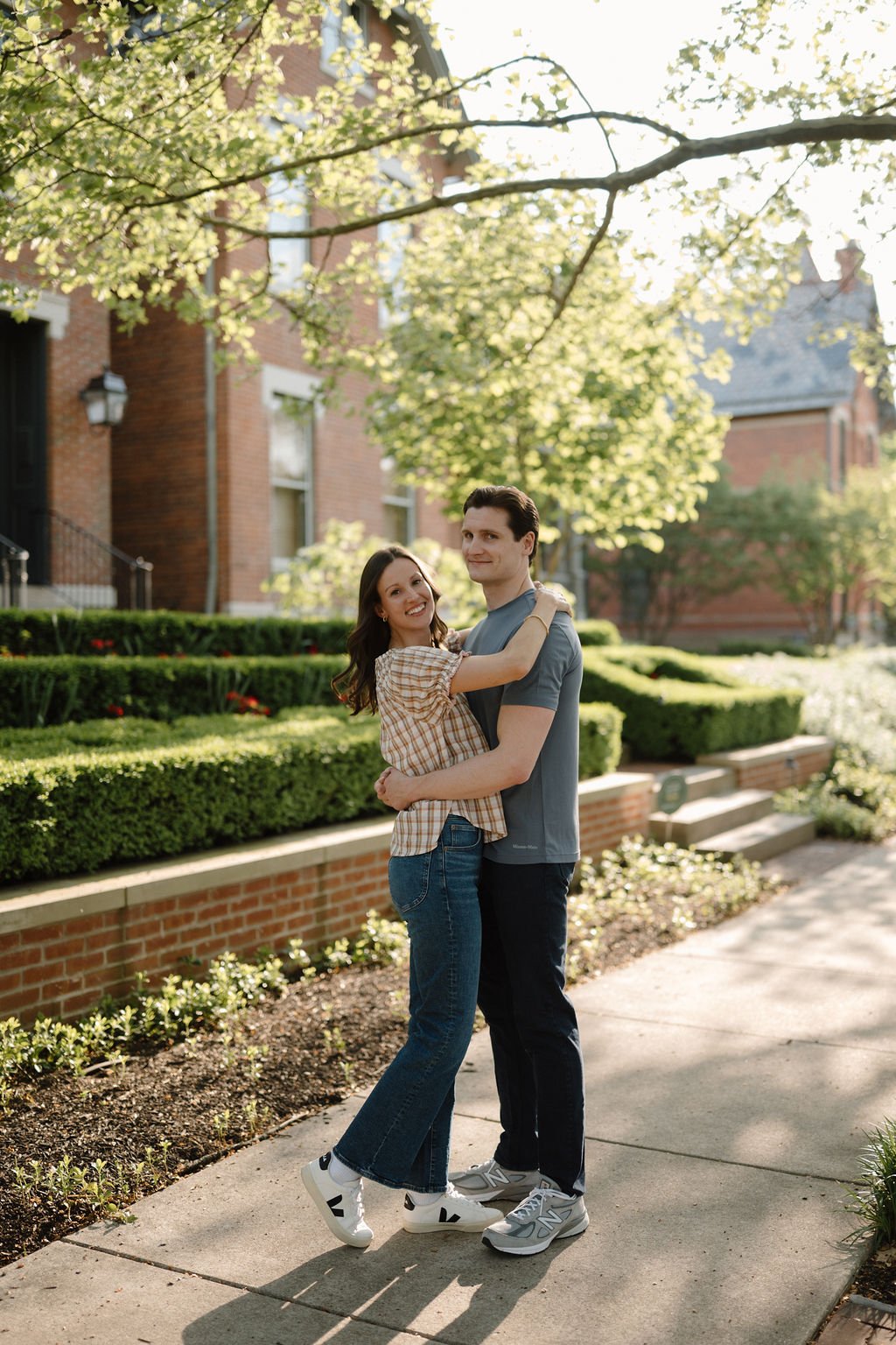 Couple hugging on a sunny sidewalk in front of a brick home with manicured hedges, a charming idea for historic district engagement photoshoot locations.