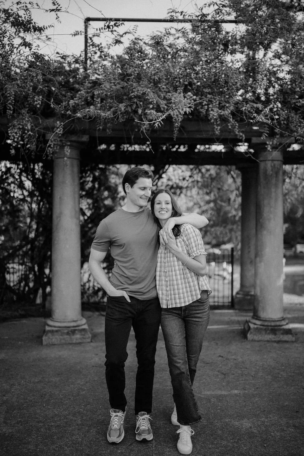 Black and white photo of a couple walking together under a vine-covered pergola, a timeless option among outdoor engagement photoshoot locations.