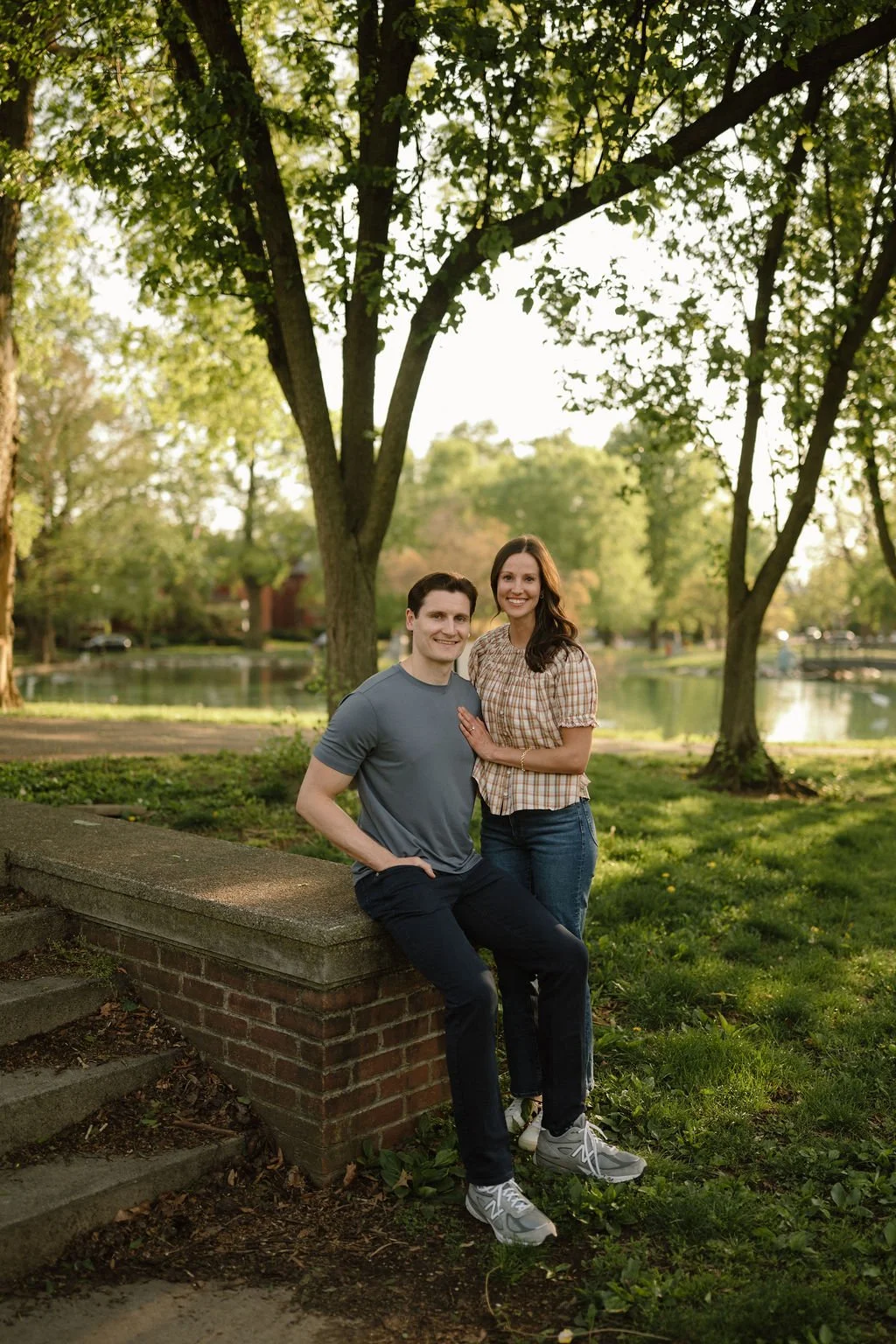 Couple posing by a brick wall in a park with a pond and trees behind them, showing how waterfront parks make great engagement photoshoot locations.