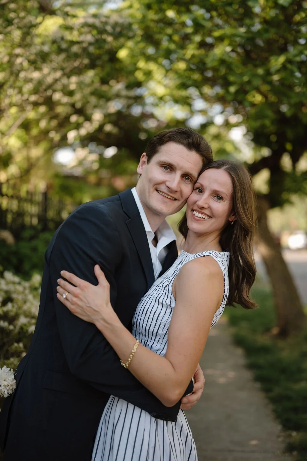 Couple hugging cheek to cheek and smiling at the camera under leafy trees, a sweet close-up for tree-lined engagement photoshoot locations.