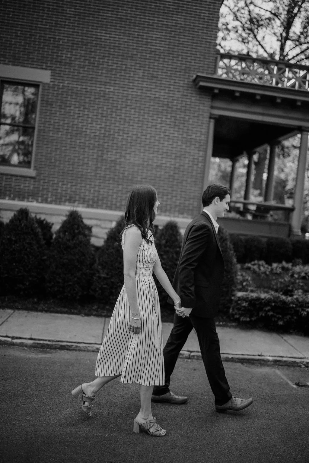 Black and white photo of a couple holding hands and walking past a historic brick home with a front porch.