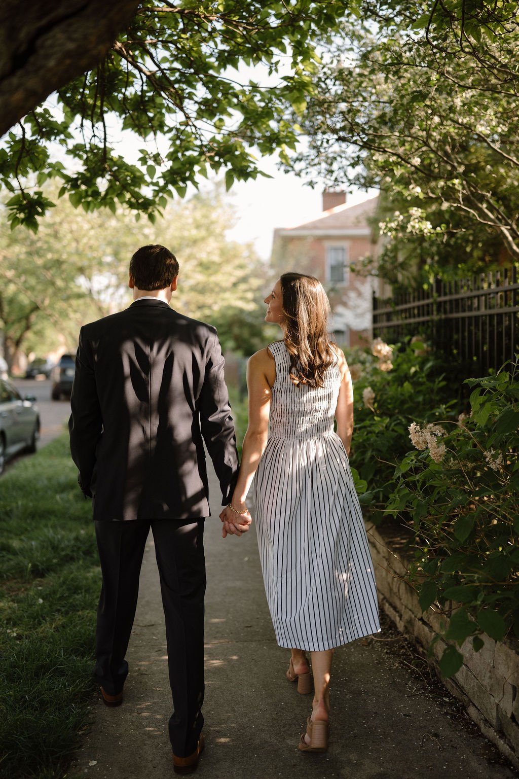Couple walking hand in hand away from the camera down a shady sidewalk toward a brick house.