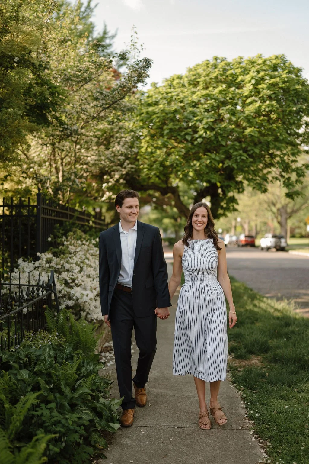 Couple holding hands on a sidewalk lined with white azaleas and wrought iron fencing, one of the classic neighborhood engagement photoshoot locations.