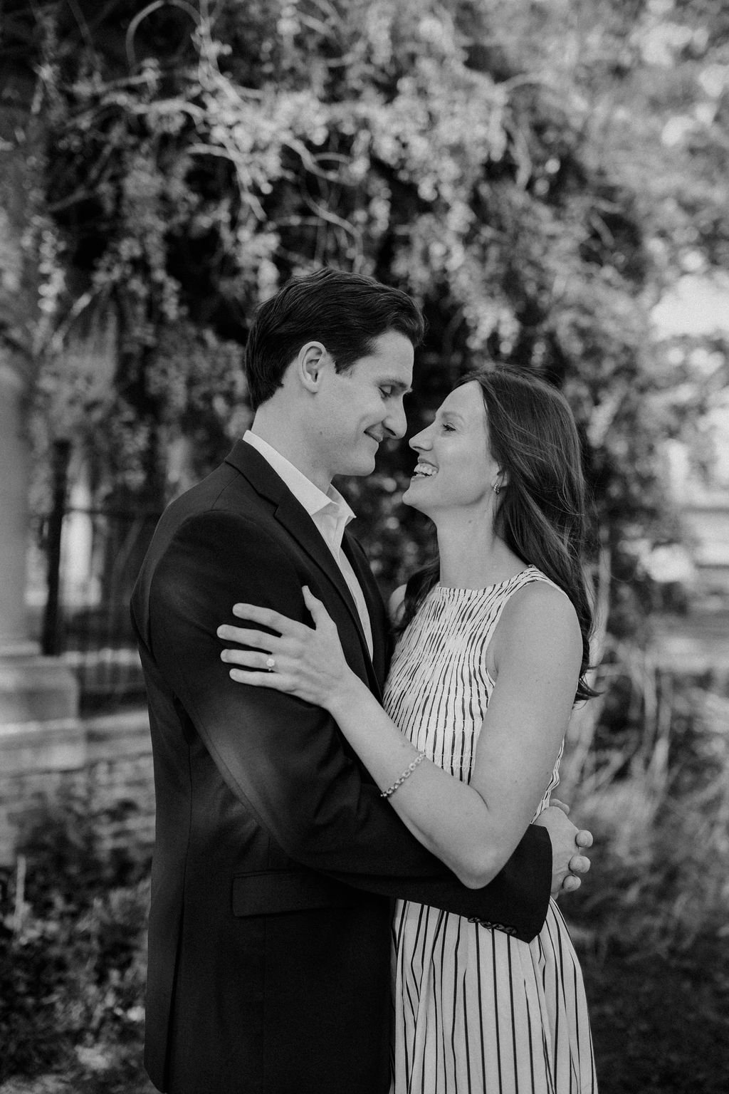 Black and white photo of a couple embracing and laughing with her ring hand resting on his suit jacket.