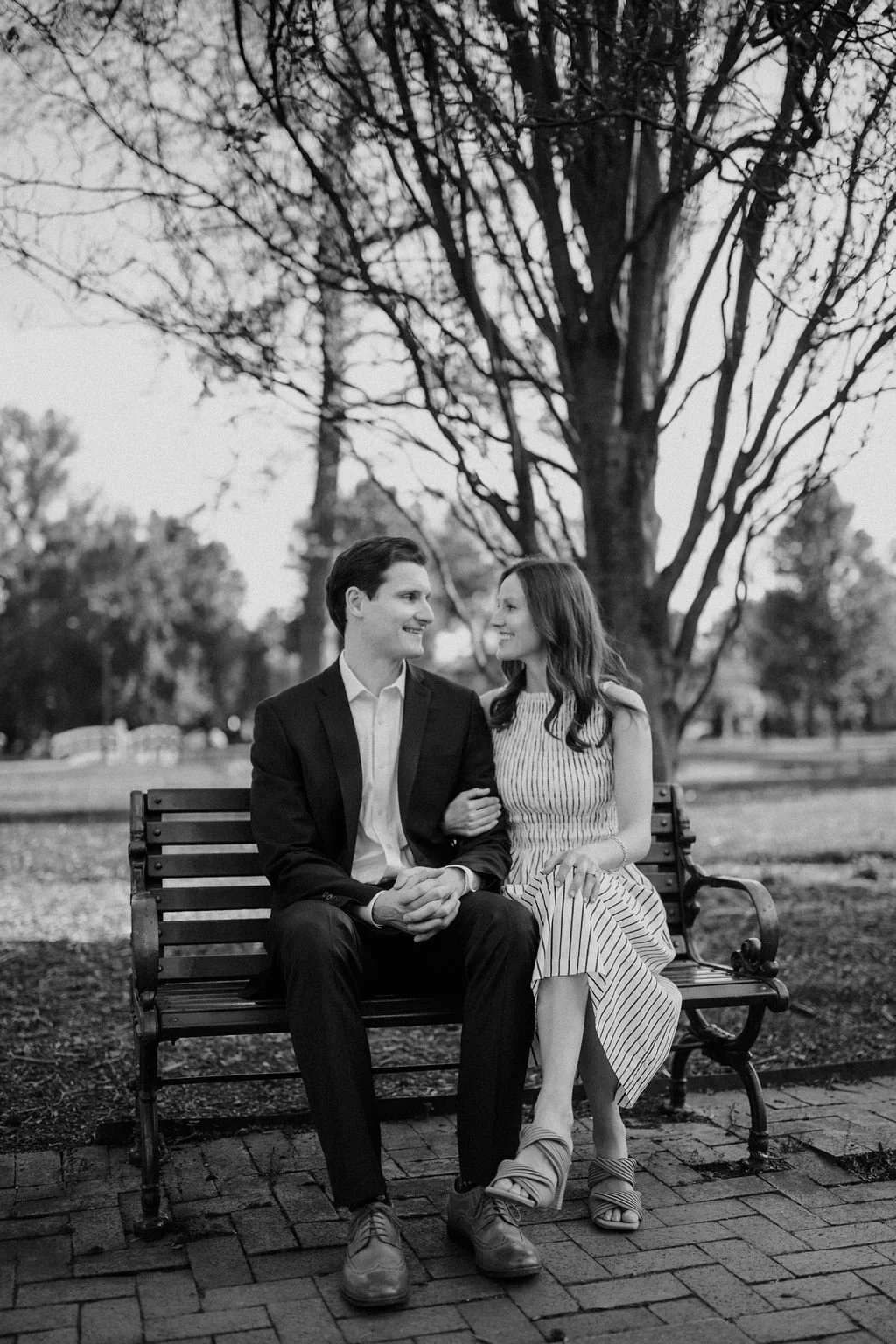 Black and white photo of a couple sitting on a park bench, smiling at each other under bare tree branches.