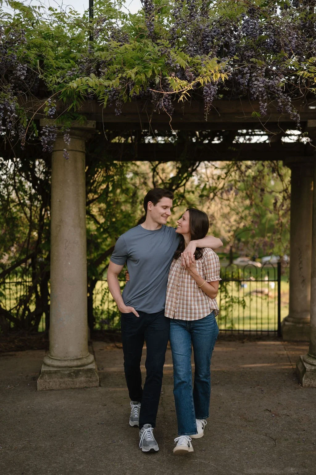 Couple strolling under a stone-column pergola covered in purple wisteria, a perfect backdrop for garden engagement photoshoot locations.
