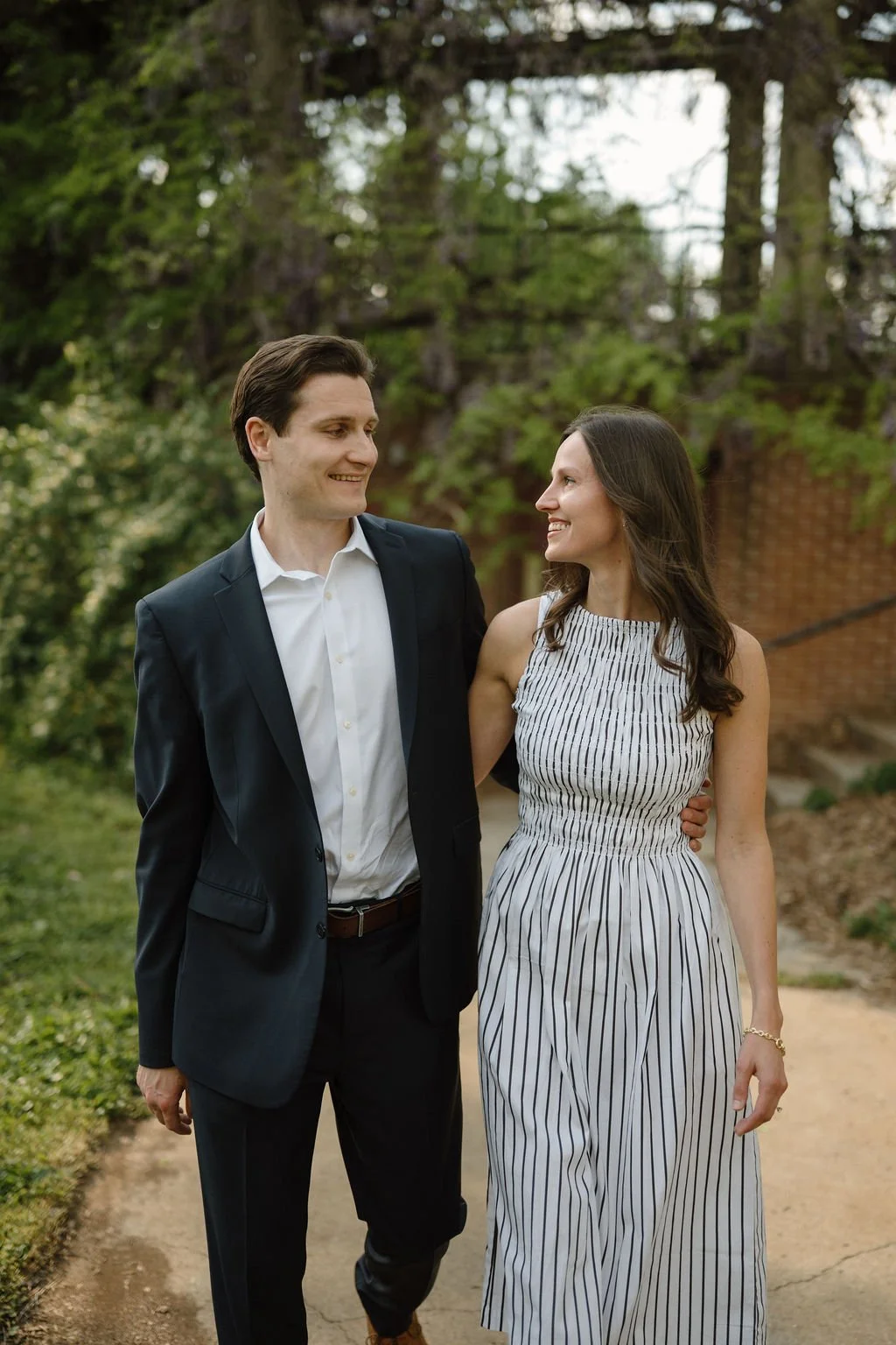 Couple walking arm in arm along a garden path beside wisteria vines and a brick wall, a lovely setting for spring engagement photoshoot locations.