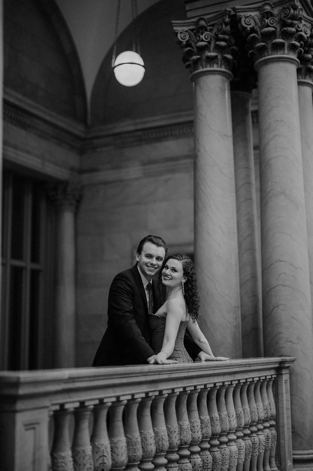 Black and white portrait of a smiling couple leaning on a marble balustrade with columns and a globe pendant light