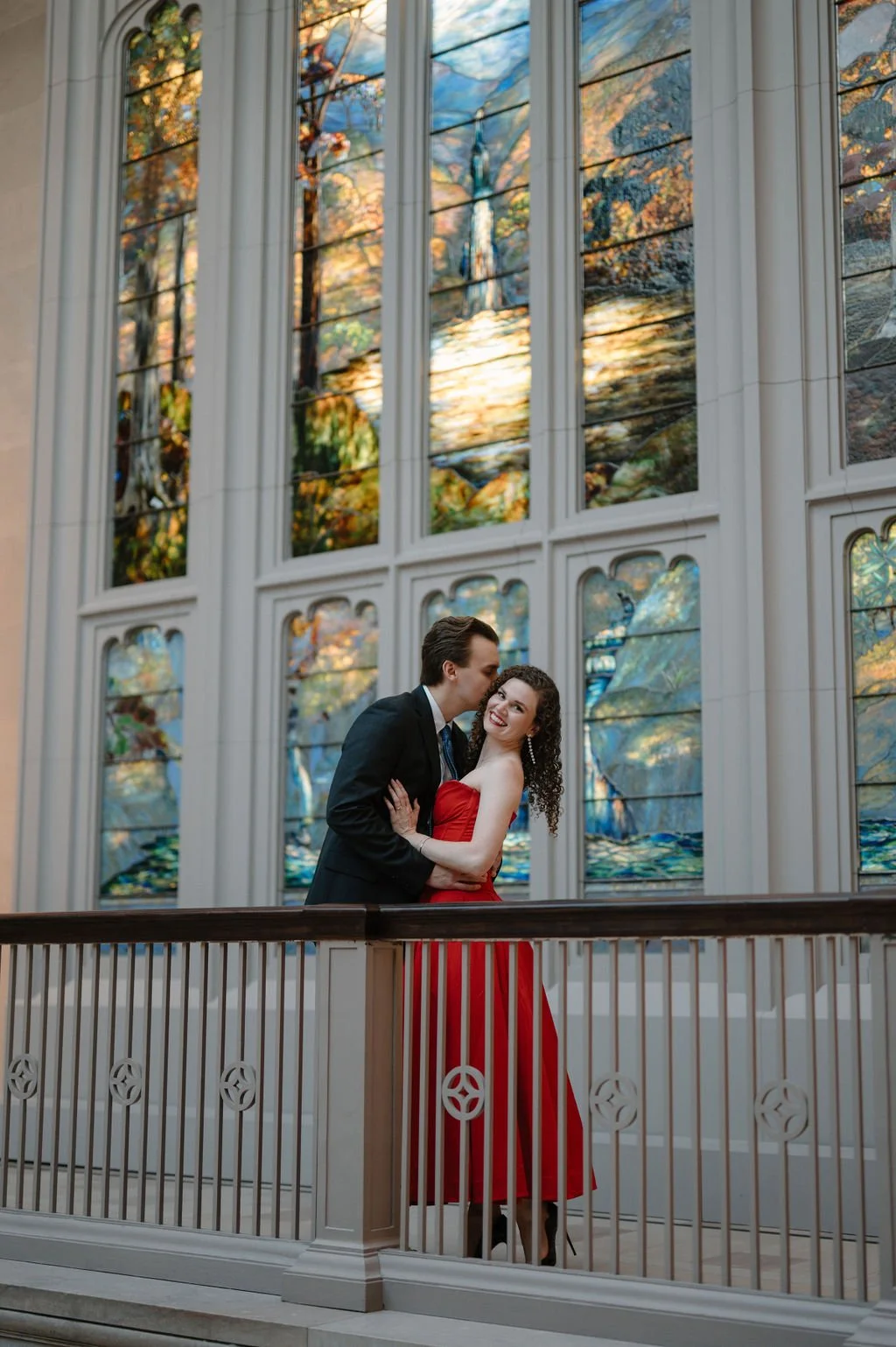 Chicago engagement photos of a couple embracing on a balcony in front of Tiffany stained glass windows as he kisses her cheek