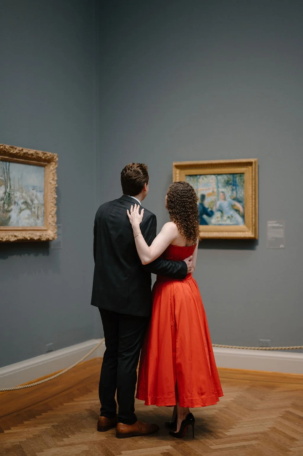 Couple seen from behind, her hand on his shoulder, admiring paintings in a blue-gray museum gallery