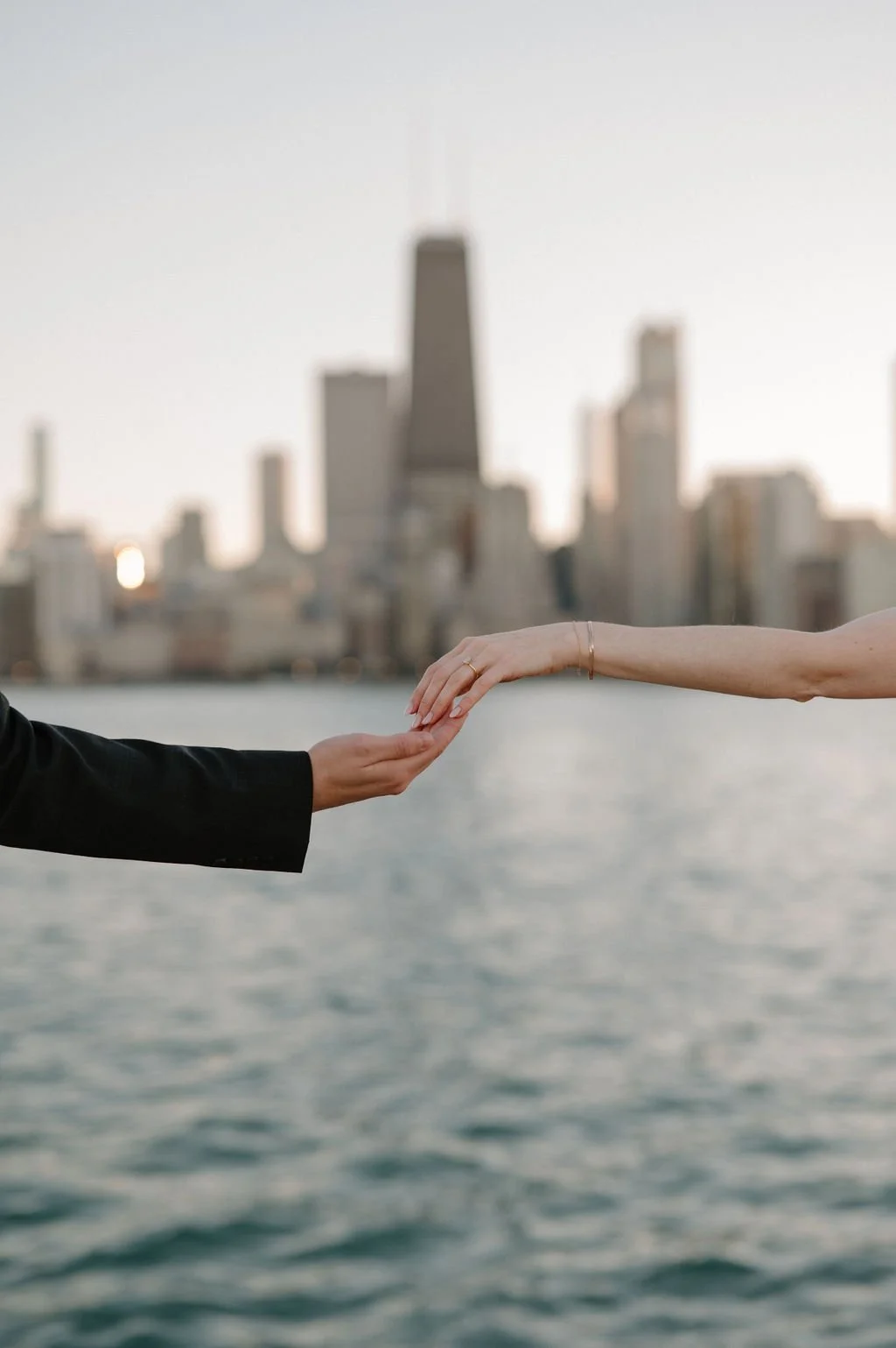 Close-up of a man's and woman's reaching hands, her engagement ring visible, with the Chicago skyline softly blurred behind