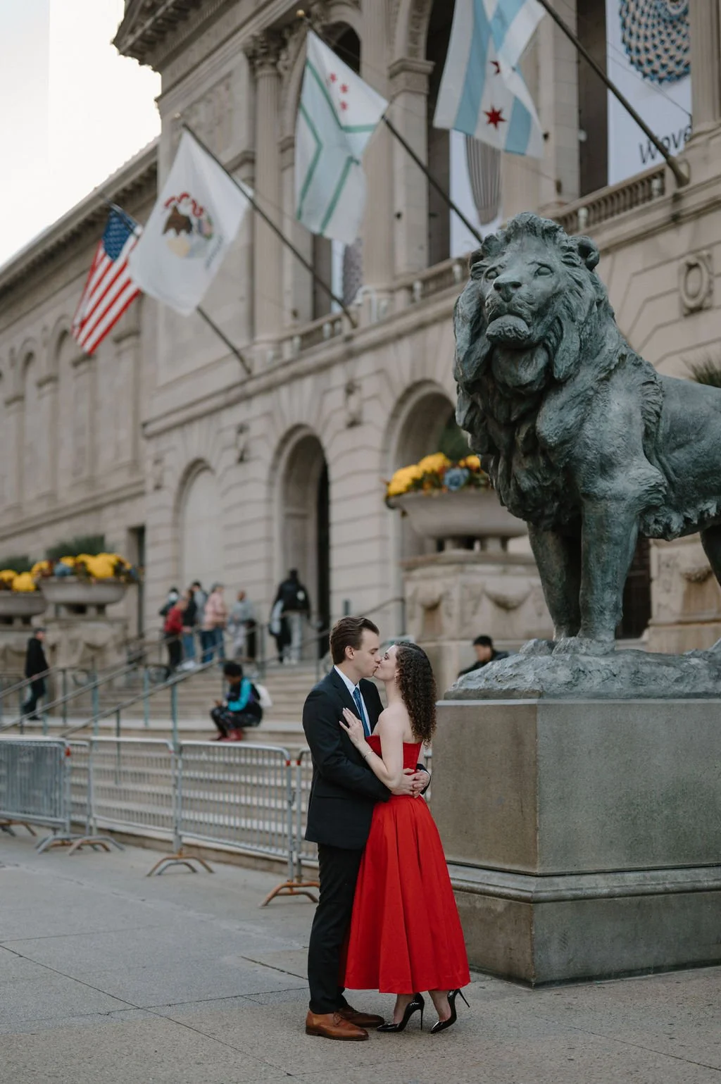 Couple kissing beside the bronze lion statue at the Art Institute of Chicago entrance, with flags overhead