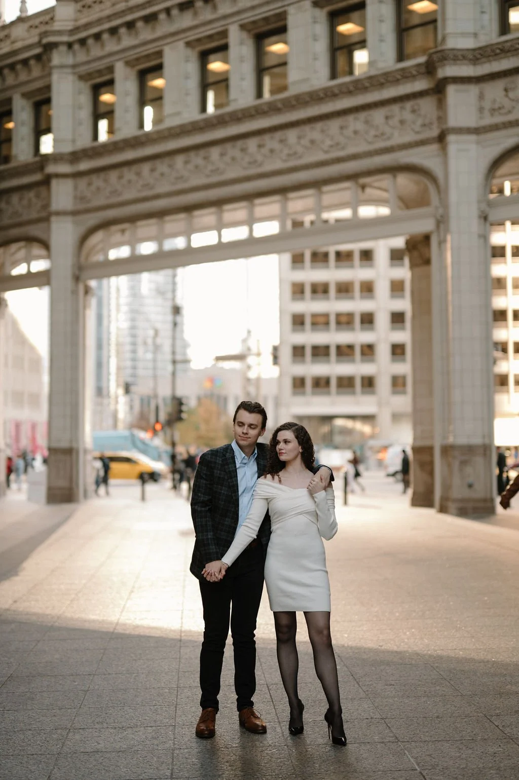 Chicago engagement photos of a couple holding hands beneath the Wrigley Building archway, her in a white dress and him in a plaid blazer