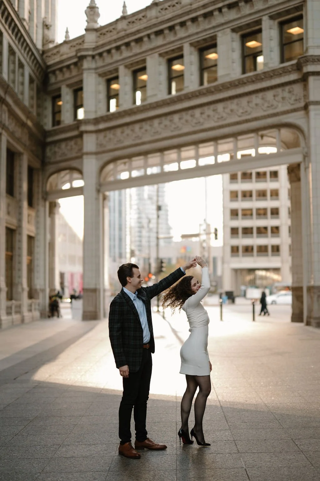 Man twirling his fiancée in a white sweater dress under the Wrigley Building archway in downtown Chicago