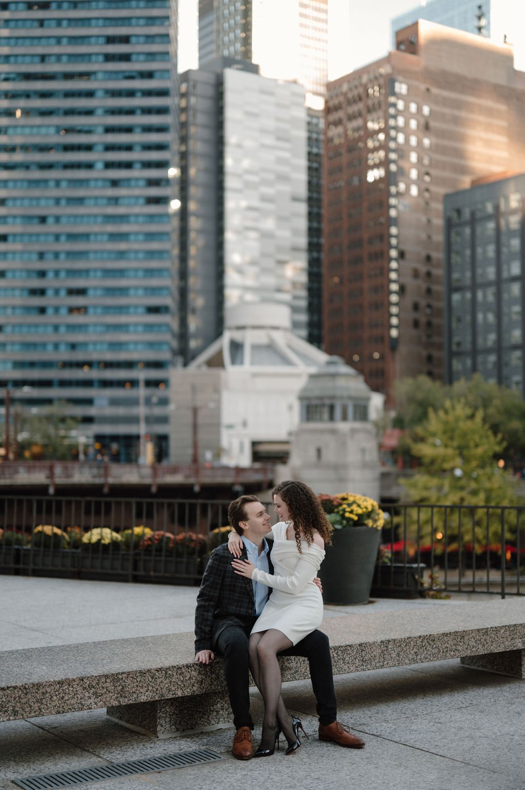 Couple sitting close on a granite bench along the Chicago Riverwalk, with fall mums and downtown skyscrapers behind them