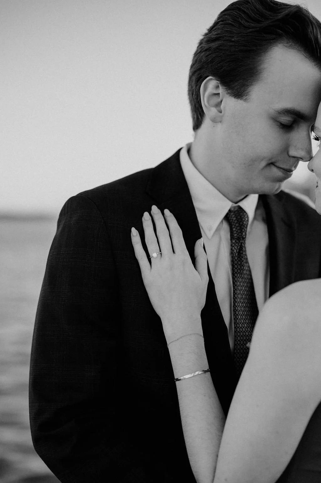 Black and white close-up of a couple touching foreheads, her hand with an engagement ring resting on his lapel