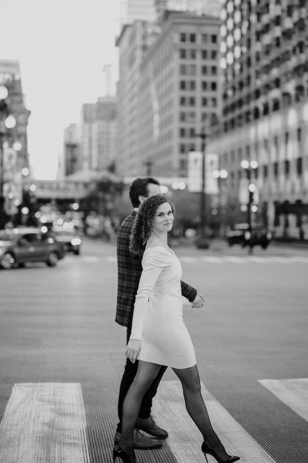 A black and white photo of an engaged couple walking hand in hand across the street during their Chicago engagement photo shoot.