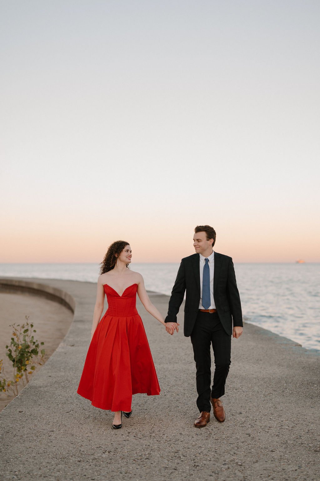 Smiling couple in a red dress and dark suit strolling hand in hand along the Chicago lakefront at golden hour
