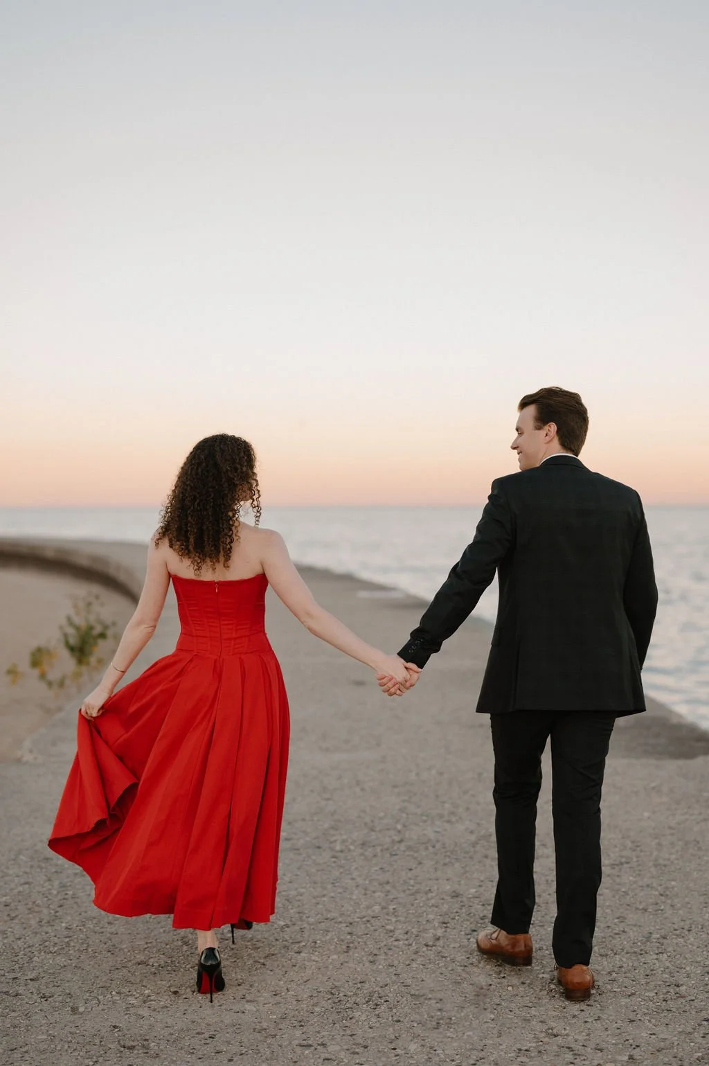 Chicago engagement photos of a couple walking hand in hand along a lakefront pier at sunset, her red skirt swaying