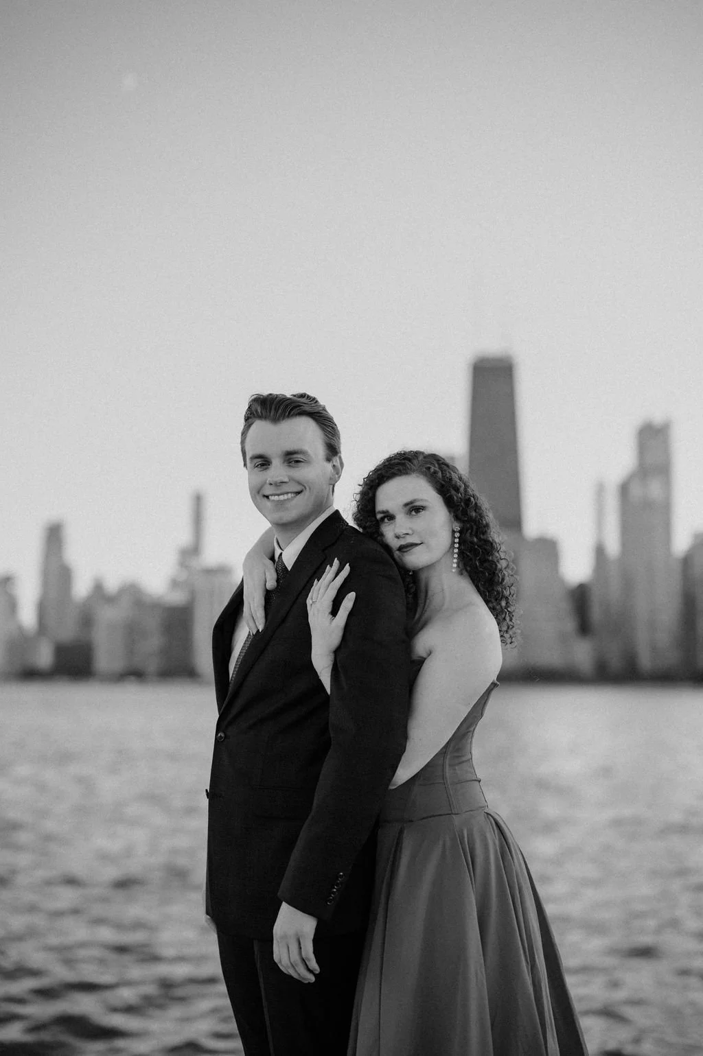 Black and white portrait of a couple by Lake Michigan, her arms around him, with the Chicago skyline blurred behind