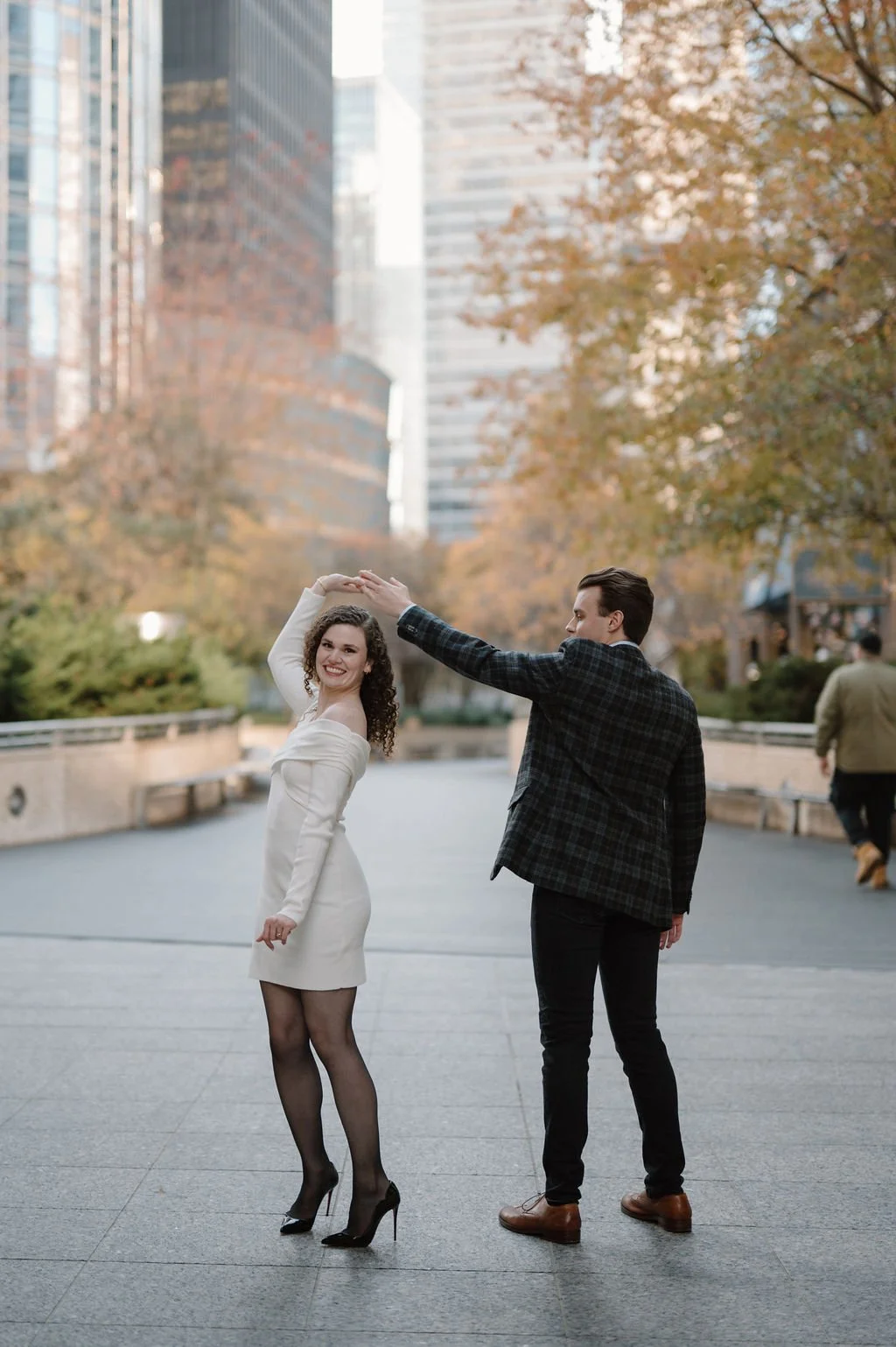 Chicago engagement photos of a woman in a white dress being twirled by her fiancé among fall trees in downtown Chicago