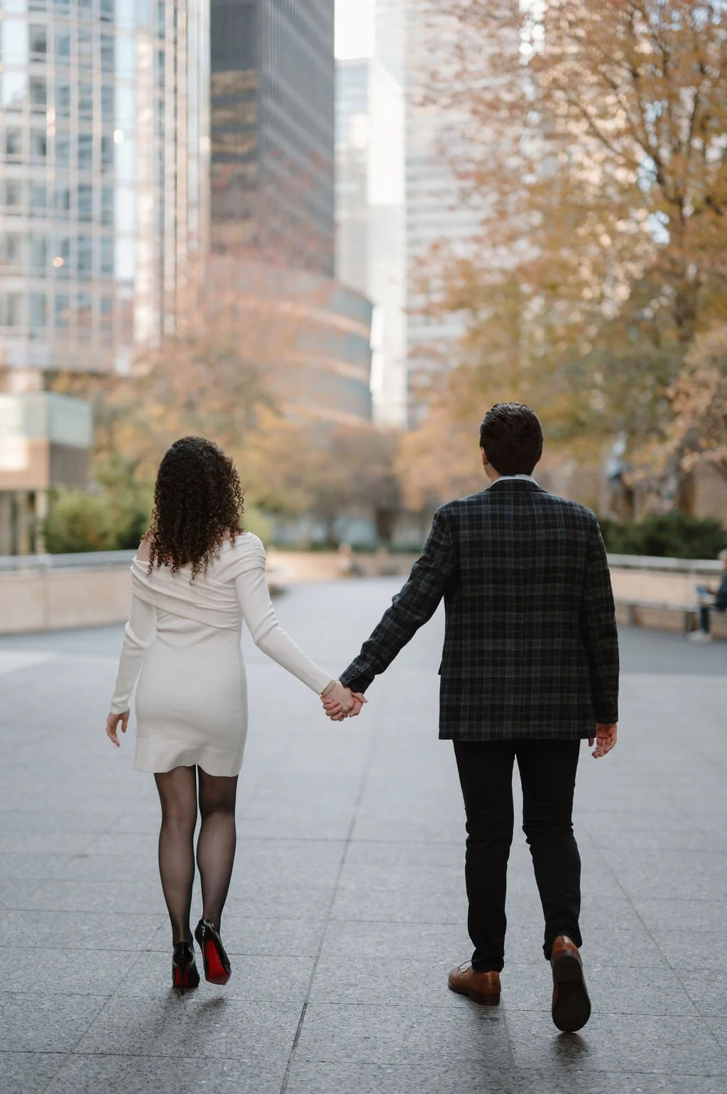 Couple holding hands and walking away from the camera through downtown Chicago, framed by fall trees and skyscrapers